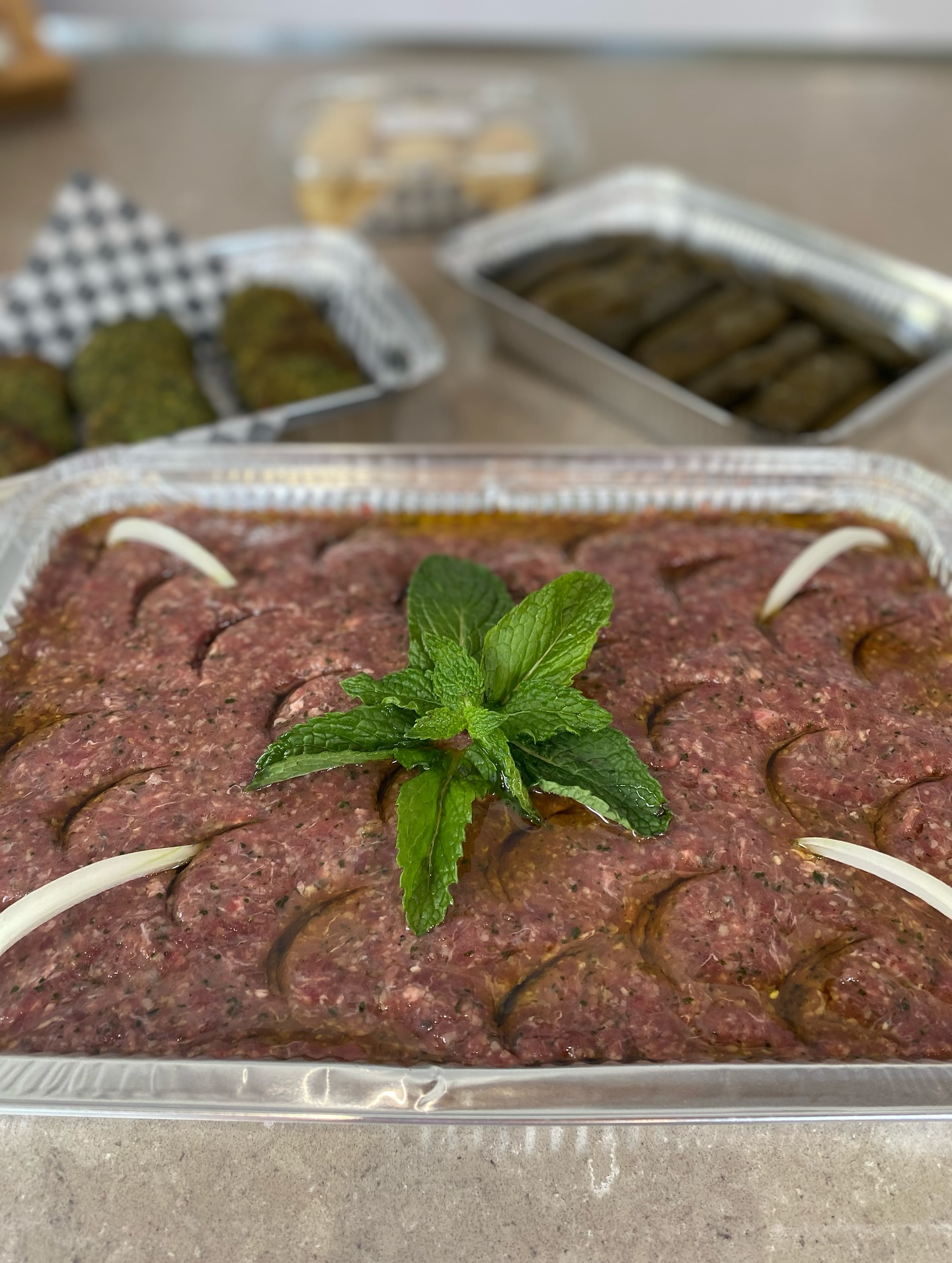 Bandeja de quibbe crudo recién preparado, con hojas de menta al centro y tiras de cebolla en las esquinas, bañado en aceite de oliva